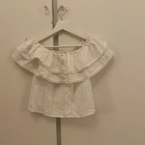 Vit offshoulder blus med volang - Supersöt vit blus med offshoulder och bred volangkant. Blusen har små bollar längs volangen och ett diskret strukturerat mönster i tyget. Perfekt för sommarens alla häng! Materialet känns lätt och luftigt. Knappt använd och jättebra kvalité