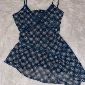 Blå asymmetrisk topp - Supersöt topp från Mexx i blått med blommigt mönster och tunna axelband. Toppen har v-ringning och är tillverkad i ett lätt, luftigt material som passar perfekt för varmare dagar. Snygg asymmetrisk nederkant och fina spetsdetaljer vid kanterna.