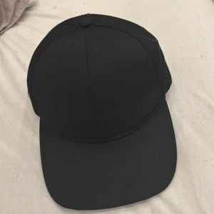 Svart keps från Prada i nylon - Snygg svart keps från Prada med klassisk böjd skärm och justerbar rem bak. Kepsen har Pradas ikoniska metallmärke på sidan och är tillverkad i slät nylon som ger en clean och modern look. Perfekt för dig som gillar exklusiva accessoarer med street vibe.