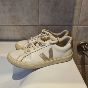 Vita Veja sneakers med gulddetaljer - Snygga vita Veja sneakers i skinn med guldfärgade detaljer på hälen och sidan. Klassisk låg modell med snörning och platt sula. Perfekta för dig som gillar stilrena och trendiga skor med en touch av metallic. 