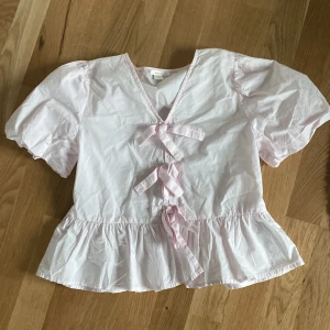 Ljusrosa blus med rosetter från Gina Tricot - Supersöt ljusrosa blus från Gina Tricot med puffiga korta ärmar, v-ringning och tre dekorativa rosetter framtill. Blusen har volangkant nedtill och är gjord i mjuk bomull. 