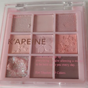 K'APEINE rosa ögonskuggspalett Korean makeup - Säljer en ögonskuggspalett med 9 nyanser i rosa, nude, brunt och glitter. Paletten har både matta och skimrande skuggor, vissa med blommönster i pressningen. Perfekt för kreativa och söta sminkningar.