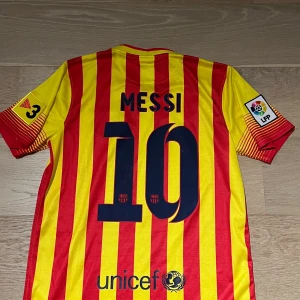 Barcelona Messi #10 randig fotbollströja - Säljer en officiell FC Barcelona fotbollströja från Nike med Messi #10 på ryggen. Tröjan är gul och röd med vertikala ränder, klubbmärke på bröstet och Qatar Airways-tryck. Tillverkad i lätt Dri-FIT-material som andas, perfekt för match eller träning.