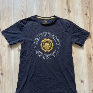 Svart t-shirt från Batistini strl M - Svart t-shirt från Batistini med tryck i vitt och gult framtill där det står 'University Campus'. Rund halsringning med gul detalj på insidan. Perfekt för en avslappnad stil.