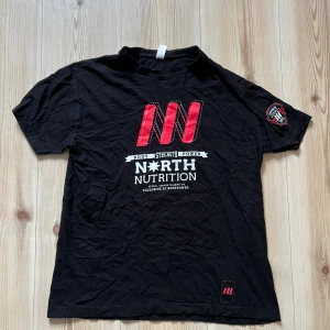 Svart North Nutrition t-shirt M - Cool svart t-shirt från North Nutrition med stort rött och vitt tryck på bröstet och patch på ärmen. Klassisk rund hals och korta ärmar. Materialet är mjuk bomull, perfekt för gymmet eller chill. Snygg detalj med logga nertill.