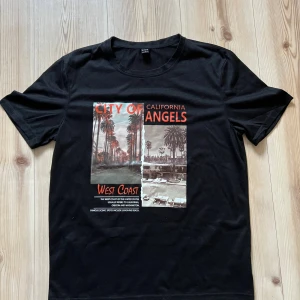Svart t-shirt från SHEIN strl L - Cool svart t-shirt från SHEIN med tryck 'City of Angels' och palmer i LA-stil på bröstet. T-shirten har rund hals och korta ärmar. Materialet är mjuk bomull som känns skönt mot huden. Perfekt för dig som gillar streetwear och snygga prints.