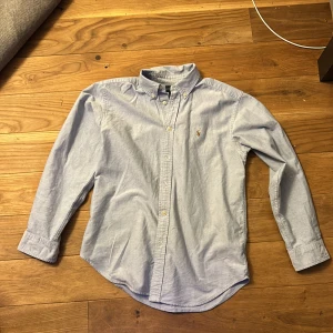 Ljusblå skjorta från Ralph Lauren - Klassisk ljusblå skjorta från Ralph Lauren med button down-krage och broderad gul logga på bröstet. Skjortan har långa ärmar, knappar framtill och är tillverkad i mjuk bomull. Perfekt för en clean och stilren look. Passar runt 160-170