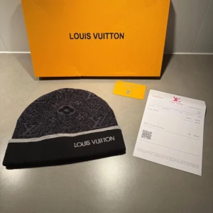 Louis Vuitton svart mönstrad mössa - Exklusiv svart mössa från Louis Vuitton med ikoniskt monogrammönster i mörkgrått. Mössan har en bred svart kant med grå rand och broderad Louis Vuitton-logga framtill. Tillverkad i mjuk ullblandning för maximal komfort och stil.