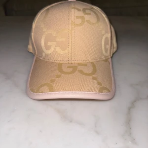 Guccci keps - Snygg beige keps från Guccci. Kepsen har böjd skärm med ljusrosa kant och justerbart spänne bak i läder. Tillverkad i bomull och polyester, perfekt för dig som vill ha en exklusiv och trendig accessoar.