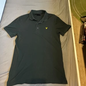 Mörkgrön pikétröja från Lyle & Scott - Snygg mörkgrön pikétröja från Lyle & Scott med klassisk krage och korta ärmar. Tröjan har det ikoniska gula fågel-loggan broderad på bröstet och är tillverkad i mjuk bomull. Perfekt för en clean och stilren look.