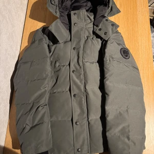 Canada Goose black label - mörkgrå pufferjacka från Canada Goose med huva och svart logotyp-patch på ärmen. Jackan har knappar och dragkedja framtill, quiltad design och är fodrad med svart innerfoder.