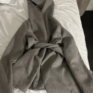 Grå omlott kappa med bälte - Säljer en stilren grå kappa med omlott-design och bälte i midjan. Jackan har lång ärm och en mjuk, lite tjockare känsla som passar perfekt när det är kyligt ute. Enkel och clean look som funkar till många outfits.