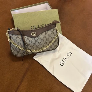 Gucci brun axelväska med kedja - Snygg axelväska från Gucci i brunt skinn och beige canvas med klassiskt GG-mönster. Väskan har guldfärgad kedja, dragkedja upptill och GG-logga i metall. Medföljer både dustbag och extra axelrem i skinn. Perfekt storlek för det viktigaste.