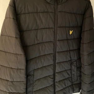 Svart pufferjacka från Lyle & Scott - Svart pufferjacka från Lyle & Scott med huva och gul broderad logga på bröstet. Jackan har horisontella quiltade paneler och dragkedja framtill. Perfekt för kalla dagar när du vill hålla stilen enkel och clean.