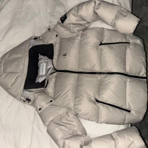 Beige pufferjacka från Calvin Klein - Säljer en varm beige pufferjacka från Calvin Klein med huva och svarta dragkedjor. Jackan har två sidofickor med dragkedja och CK-logga på bröstet. Perfekt för kalla dagar och har en modern, clean look. Köpte den har it använt den mer än en vecka så den är helt ny.