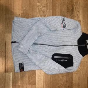 Ljusgrå Sail Racing kofta - Ljusgrå ribbad kofta från Sail Racing, Antarctica Expedition edition. Tröjan har dragkedja nedifrån och bakifrån och svart bröstficka med dragkedja. Snygga detaljer med patch på nederkanten och tryck på ärmen. Glöm inte att Sail Racing är ett känt klädesmärke!