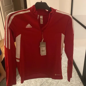 Röd Adidas långärmad träningströja - Säljer en röd långärmad träningströja från Adidas med vita detaljer och klassiska ränder på axlarna. Tröjan har half zip framtill och är tillverkad i polyester med Aeroready-teknologi. Perfekt för sport eller chill. Adidas-logga på bröstet.