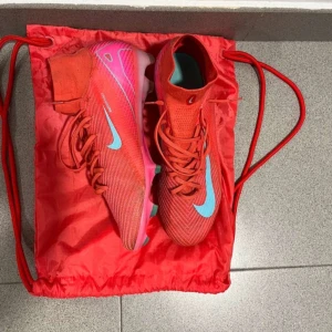 Nike Mercurial röda fotbollsskor - Säljer ett par Nike Mercurial fotbollsskor i röd och rosa med ljusblå swoosh. Skorna har snörning, hög strumpliknande krage och mönstrad sula för extra grepp. Ovandelen är i syntetmaterial och designen är riktigt sportig och modern.
