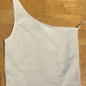 Vit oneshoulder ribbad topp Gina Tricot - Säljer en vit ribbad topp från Gina Tricot i storlek S. Modellen är oneshoulder med en axelbandslös sida, vilket ger en snygg och modern look. Materialet är mjukt och stretchigt, perfekt för att skapa en cool stil. Passar dig som gillar trendiga detaljer.