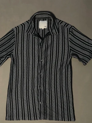 Blå och vit randig skjorta - Snygg skjorta med blå och vita vertikala ränder. Skjortan har korta ärmar, klassisk krage och knäppning framtill. Perfekt för dig som gillar retrostil och vill sticka ut lite extra.