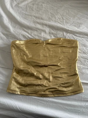 Guldig glittrig tubtopp - Snygg guldig tubtopp med glittrig finish. Toppen är axelbandslös och har en enkel, rak passform som sitter tight mot kroppen.