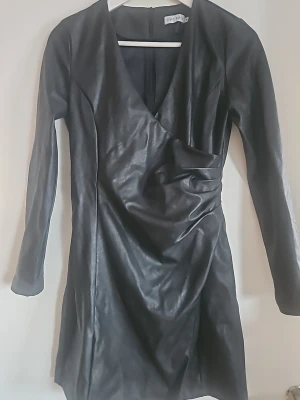 Svart omlottklänning i skinnimitation - Snygg svart kort omlottklänning i skinnimitation. Klänningen har v-ringning och långa ärmar, samt en draperad detalj framtill som ger en cool look. Perfekt för dig som vill sticka ut med en edgy stil.