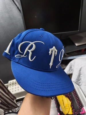 Blå snapback med broderad text - Snygg blå snapback-keps med stor, vit broderad text runt hela kepsen. Klassisk platt skärm och justerbar baksida. Perfekt för dig som vill sticka ut med en streetwear-look. Materialet är bomullstyg och kepsen har en modern, ungdomlig vibe.