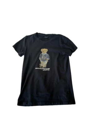 Mörkblå t-shirt med björntryck från Polo Ralph Lauren - Mörkblå t-shirt från Polo Ralph Lauren med ikoniskt björntryck på bröstet. Klassisk passform med rund hals och korta ärmar. Perfekt för en avslappnad och cool stil. Bara att höra av sig vid frågor!