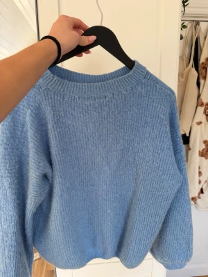 Ljusblå stickad tröja med rund hals - Mysig ljusblå stickad tröja med rund halsringning och ribbade muddar. Tröjan har en avslappnad passform och är perfekt för lager-på-lager. Materialet känns mjukt och fluffigt, vilket gör den extra skön att bära under kyliga dagar.
