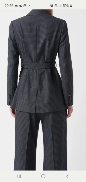 Mörkgrå kavaj med bälte från Studio - Snygg mörkgrå kavaj med diskret rutmönster och tillhörande midjebälte från Studio Collection Project. Klassisk krage, markerad midja och långa ärmar med knappar vid ärmslut. Perfekt för en stilren och modern look.