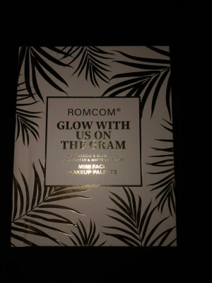 ROMCOM Glow With Us On The Gram Palette - Ögonskuggspalett från ROMCOM med nio nyanser i både matta och skimrande toner. Färgerna går i nude, rosa, lila och brons med både neutrala och glittriga skuggor. Paletten har ett stilrent vitt fodral med svarta palmblad och gulddetaljer.