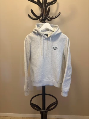 Grå hoodie från A.P.C. Paris - Snygg ljusgrå hoodie från A.P.C. med broderad svart logga 'A.P.C. RUE MADAME PARIS' på bröstet. Klassisk modell med huva och dragsnören, ribbade muddar vid ärmar och nederkant. Tillverkad i mjuk bomullsblandning för skön känsla. Ny pris 3200kr mitt pris 1199, priset är inte hugget i sten. använd vid 2 tillfällen 