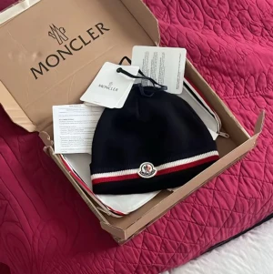 Svart Moncler mössa med logga - Snygg svart mössa från Moncler med klassisk logga framtill och vit-röd rand längs kanten. Mössan är ribbstickad och har en stilren, tight passform. Perfekt accessoar för att lyfta din outfit under kyliga dagar.