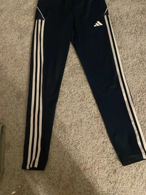 Svarta Adidas träningsbyxor 152 - Svarta träningsbyxor från Adidas med klassiska vita ränder längs sidorna och Adidas-logga på låret. Byxorna har resår i midjan och dragsko för bästa passform. Perfekta för träning eller chill. Materialet är mjukt och stretchigt.