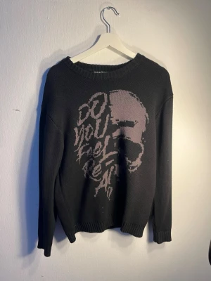 Svart stickad tröja Koroshi S - Svart stickad tröja från Koroshi med rund hals och ribbade muddar. Framsidan har ett stort grafiskt tryck i grått med texten 'Do you feel real?'. Otroligt bra kvalité med tjockare material.