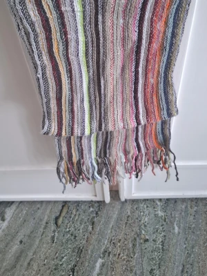 Randig halsduk med fransar - Färgglad halsduk med smala ränder i vitt, svart, grått, grönt, rött, orange och blått. Halsduken har fransar i ändarna och är tillverkad i ett mjukt, vävt material. Perfekt för att addera lite färg till din outfit.