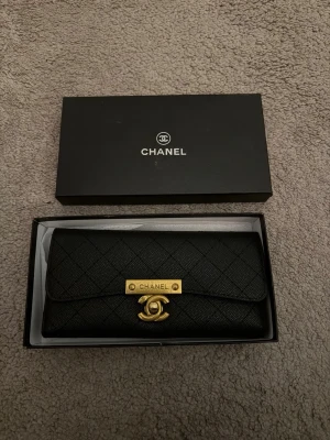 Svart plånbok  - Lyxig svart plånbok i quiltat läder med guldfärgad logga och metallspänne framtill. Klassisk rektangulär form och elegant design,  