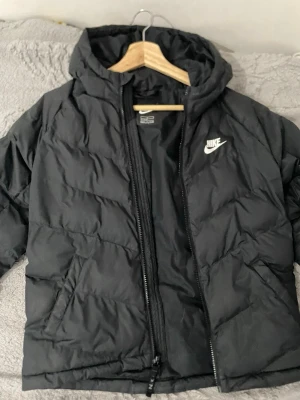 Svart dunjacka med huva från Nike - Snygg svart dunjacka från Nike med huva och dragkedja framtill. Jackan har en klassisk puffer-design och Nike-logga i vitt på bröstet. Perfekt för kalla dagar och enkel att matcha med olika outfits. Tillverkad i slitstarkt material och har långa ärmar.