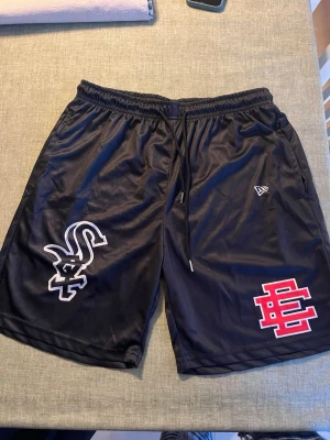 Svarta sportiga shorts med tryck - Svarta sportshorts från New Era med elastisk midja och dragsko. Ena benet har en vit Chicago White Sox-logga och det andra ett rött och vitt tryck. Tillverkade i ett glansigt, lätt polyestermaterial som andas. Perfekta för träning eller chill.