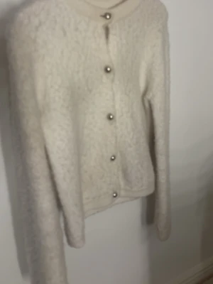 Krämvit fluffig kofta med pärlknappar - Mysig krämvit kofta med fluffig textur och stora pärlknappar framtill. Långärmad modell med rund halsringning och rak passform. Perfekt för dig som gillar en enkel men ändå elegant look. inga tecken på användning 