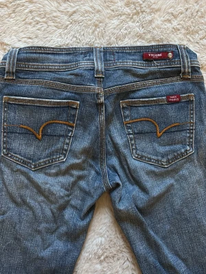 Blå bootcut jeans från Vigoss - Ett par klassiska blå bootcut jeans från Vigoss med snygga kontrastsömmar på bakfickorna och diskret tvättad look. Jeansen har fem fickor och normalhög midja. Perfekta för en avslappnad och trendig stil. En knapp saknas.