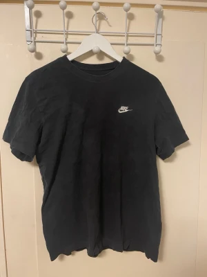Svart Nike T-shirt med logga - Svart T-shirt från Nike med klassisk vit logga på bröstet. Modellen är enkel med rund halsringning och korta ärmar. Perfekt för en avslappnad och sportig stil. Tillverkad i mjukt bomullsmaterial för skön komfort.