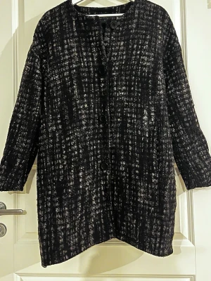 Svartvit/grå mönstrad ullkappa - Stilren svartvit kappa med rutigt mönster och stora knappar framtill. Rak passform och långa ärmar, perfekt för kyligare dagar. Materialet är ull och polyester, varm, mjuk och lite fluffig, vilket ger en mysig känsla. Enkel och trendig design som passar till många outfits.