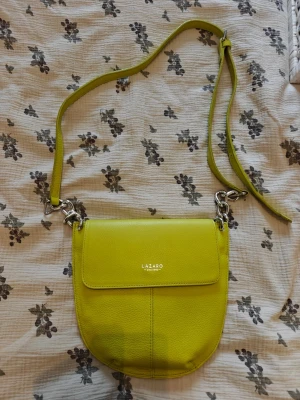 Limegrön axelväska  - Snygg limegrön axelväska från Lazaro med justerbar rem och silverfärgade metalldetaljer. Väskan har en rundad form och är tillverkad i strukturerat läder. Perfekt för dig som vill sticka ut med en färgglad accessoar. Läder. Från märket Lazaro i Argentina, se alla bilder för detaljer, finns lite slitage på bandet, torr.  Ingår en spegel.
