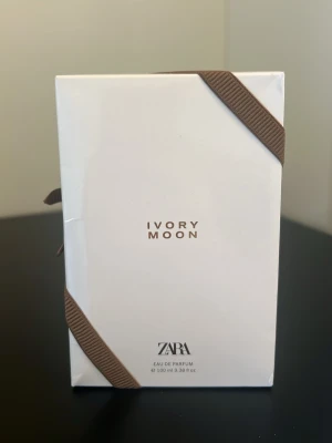 ZARA Ivory Moon Eau de Parfum 100ml - ZARA Ivory Moon Eau de Parfum i en stilren vit förpackning med bruna banddetaljer. Flaskan rymmer 100 ml och har en minimalistisk design med guldtext. Perfekt för dig som gillar eleganta och moderna dofter.