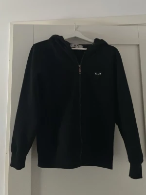 Svart hoodie från Comme des Garçons - Svart hoodie med dragkedja från Comme des Garçons PLAY. Klassisk design med huva och det ikoniska hjärta-med-ögon-loggan broderad på bröstet. Perfekt för en clean och stilren look.