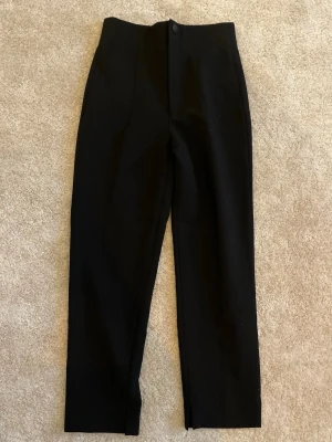 Svarta raka kostymbyxor - Stilrena svarta kostymbyxor med rak passform och pressveck. Byxorna har hög midja, dold dragkedja och knapp framtill. Perfekta för en clean och klassisk look. Materialet ser ut att vara en mjuk och lite stretchig mix.