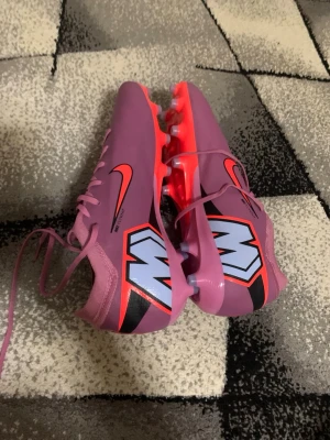 Lila Nike Mercurial fotbollsskor vapor 16 - Säljer ett par lila Nike Mercurial fotbollsskor med stora vita och svarta M-loggor på hälen och orange detaljer. Skorna har snörning och dobbar i plast för spel på gräs. Snygg och modern design som sticker ut på planen. Storlek 44.5. Jag har använt dom 1 träning på grund av att dom var stora. Pris: 1200kr