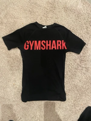  Gymshark muscleshirt  - Säljer en svart t-shirt från Gymshark med stor röd logga över bröstet. Tight passform som sitter snyggt mot kroppen och korta ärmar. Perfekt för gymmet eller när du vill ha en sportig look. Gjord i mjukt och stretchigt material.