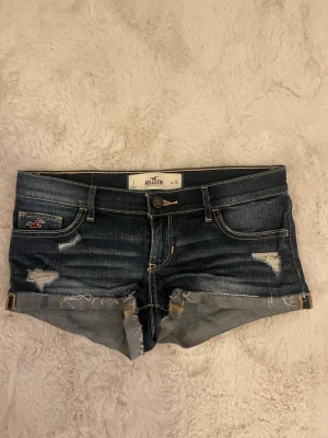 Mörkblå jeansshorts från Hollister - Snygga mörkblå jeansshorts från Hollister med slitna detaljer och uppvikta benslut. Klassisk femficksmodell med gul kontrastsöm och logga på fickan. Perfekta för varma dagar och enkel att matcha med olika toppar. W.25💕